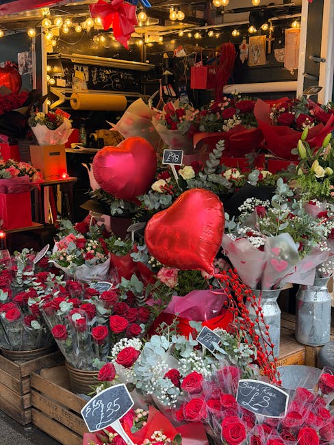An indoor display of various floral arrangements featuring red roses, white flowers, and greenery, accented by large red heart-shaped balloons. The flowers are wrapped in pink and white paper, some in
