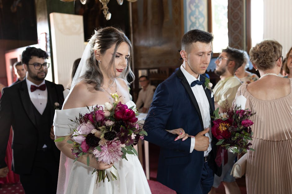 In a wedding venue with ornate wooden walls and soft natural lighting, a bride dressed in an off-the-shoulder white gown with a veil holds a vibrant floral bouquet featuring deep pink peonies, blush r