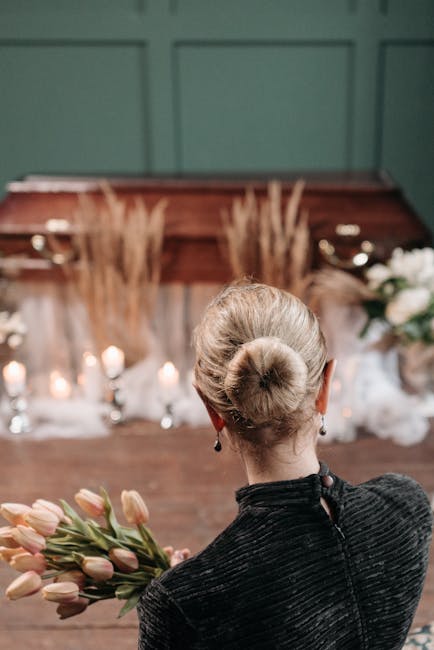 A woman with platinum blonde hair styled in an elegant bun, dressed in a dark textured top, is holding a bouquet of pale pink and peach tulips with green stems. She appears to be standing indoors in f