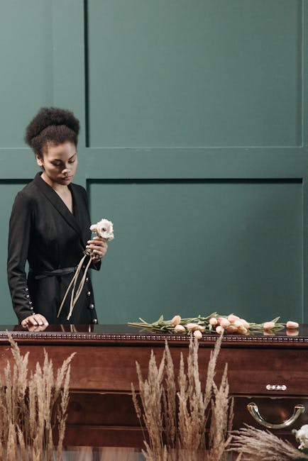 A woman dressed in black holds a small white floral arrangement in her left hand, standing beside a wooden casket that is decorated with a variety of pale pink and peach roses, tulips, and greenery. T