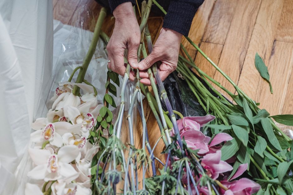 A pair of hands are arranging a floral bouquet on a wooden surface, with various fresh flowers including white orchids with pink markings and pink lilies, alongside green foliage. The flowers are held