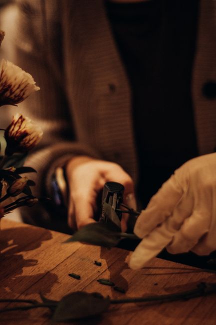 Close-up of a florist’s hands arranging a floral bouquet on a wooden surface, with a pair of white gloves and floral scissors visible. The flowers include soft pink and cream-colored blooms, accompa