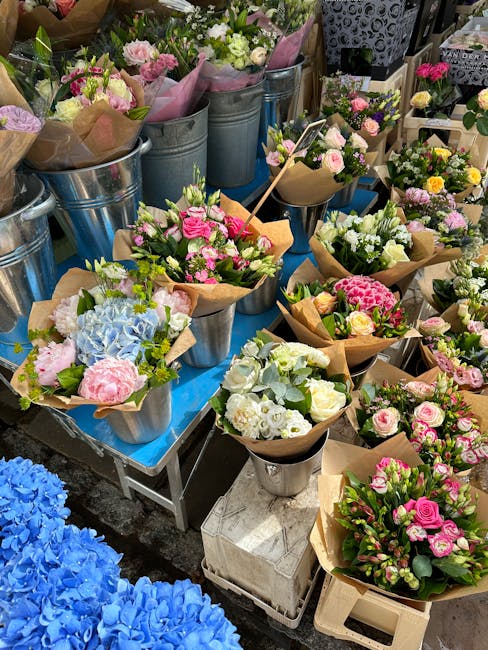 A variety of fresh floral bouquets arranged in metal buckets and wrapped in brown and pink paper, featuring pink roses, white and green carnations, and blue hydrangeas, displayed on a blue table at a