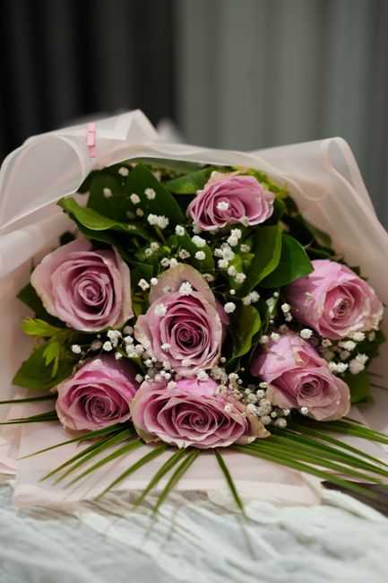 A bouquet of fresh, light pink roses with tightly layered petals, accented by delicate white baby's breath and lush green leaves. The flowers are arranged in a rounded, compact style and wrapped in so